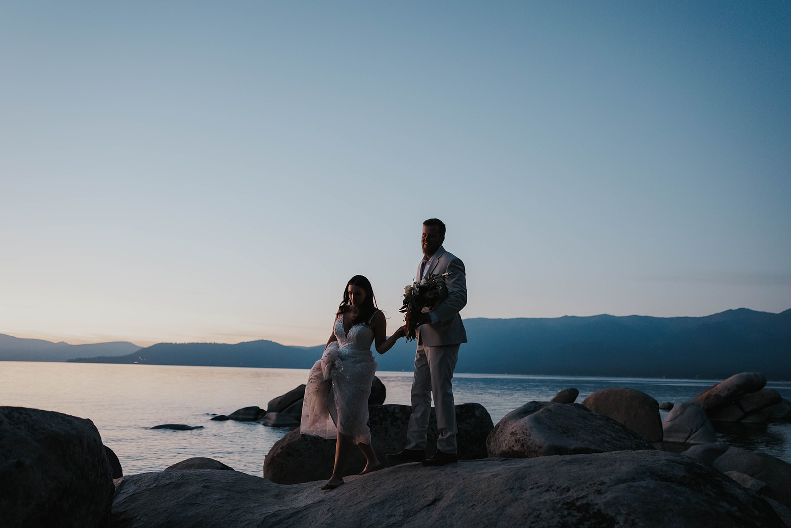 Husband and wife walk along rocks at Lake Tahoe during blue hour. 